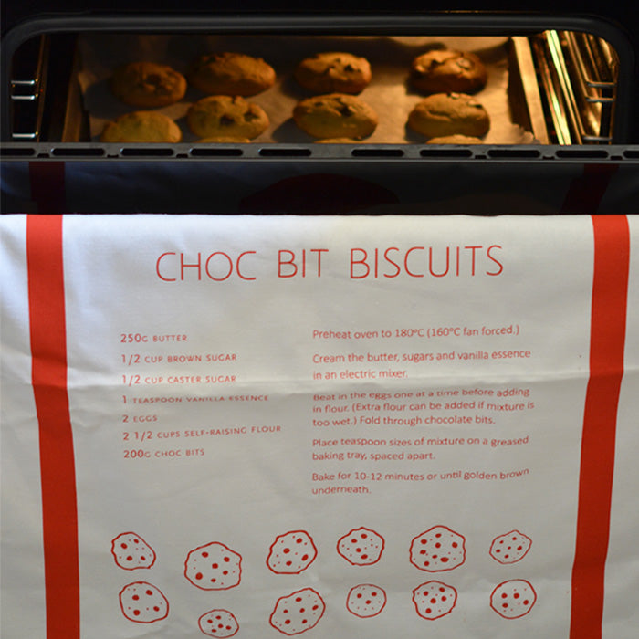 A tea towel on the rung of an over door that is open slightly to show biscuits baking. The tea towl has a recipe, thick red board and illustrated biscuits on it