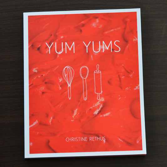 Yum Yums recipe book with a red cover, text reading Yum Yums Christine Rethus on a red cake mixture photo with illustrations of a wooden spoon, rolling pin and whisk in white on a dark wood background