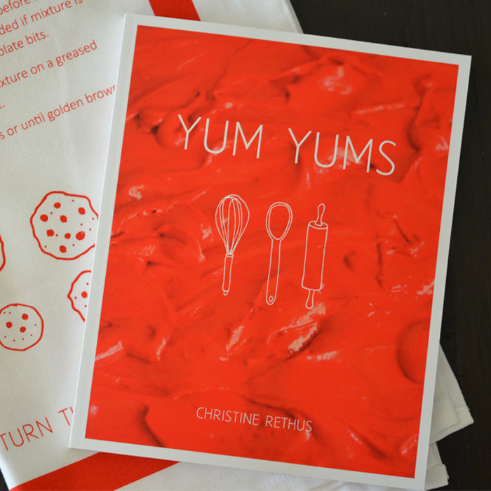 Picture of Yum Yums recipe book against a tea towel with illustrated cookies and a recipe against a dark wood background